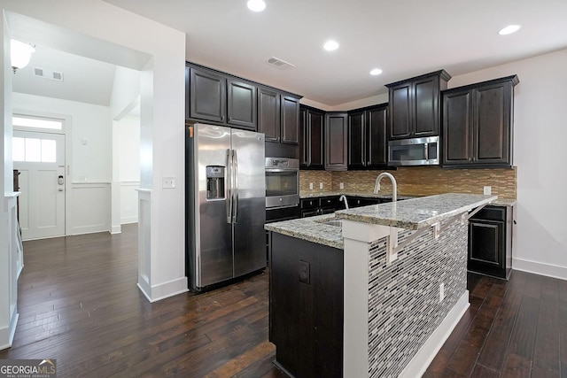 kitchen with appliances with stainless steel finishes, backsplash, light stone counters, a kitchen island with sink, and dark wood-type flooring