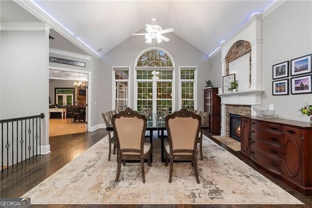 dining space featuring ceiling fan with notable chandelier, lofted ceiling, dark wood-type flooring, a stone fireplace, and crown molding