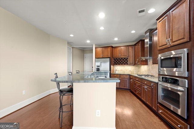 kitchen featuring stainless steel appliances, wall chimney range hood, light hardwood / wood-style flooring, a kitchen bar, and a center island with sink