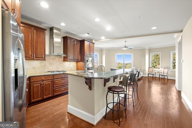 kitchen with a breakfast bar, a center island with sink, hardwood / wood-style flooring, wall chimney exhaust hood, and appliances with stainless steel finishes