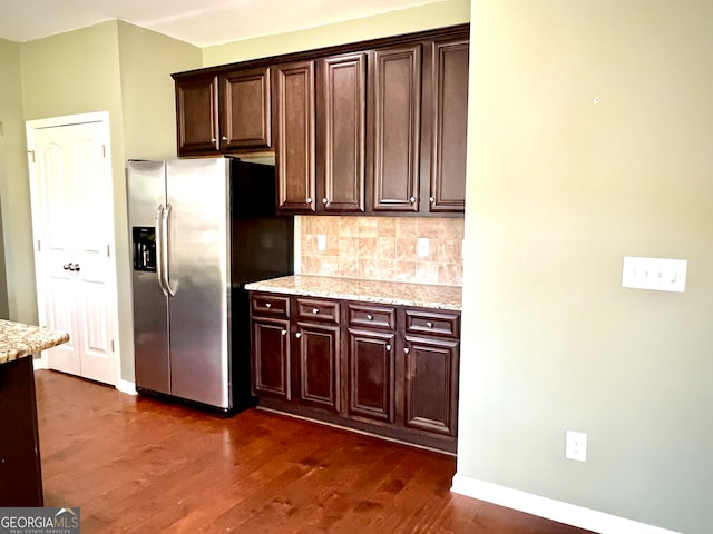 kitchen with light stone countertops, stainless steel refrigerator with ice dispenser, decorative backsplash, dark brown cabinets, and dark wood-type flooring