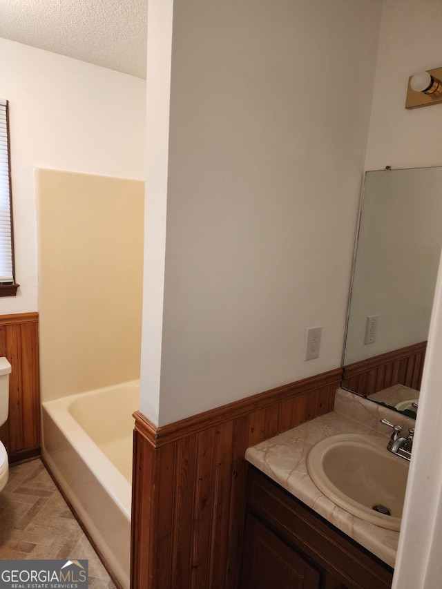 full bath with a wainscoted wall, wood walls, toilet, a bath, and a textured ceiling