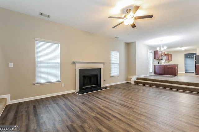 unfurnished living room featuring ceiling fan with notable chandelier, a wealth of natural light, and dark wood-type flooring