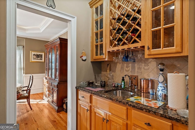 bar with decorative backsplash, light wood-type flooring, dark stone counters, a raised ceiling, and sink