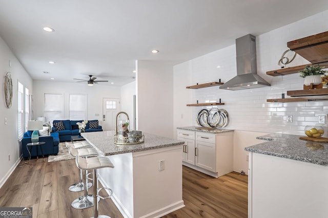 kitchen with light stone countertops, wall chimney range hood, light hardwood / wood-style flooring, a center island with sink, and white cabinets