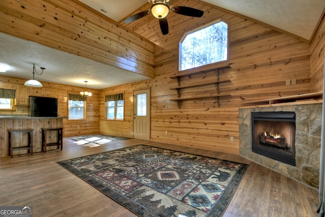 living room with a stone fireplace, dark hardwood / wood-style floors, wood walls, a textured ceiling, and ceiling fan with notable chandelier