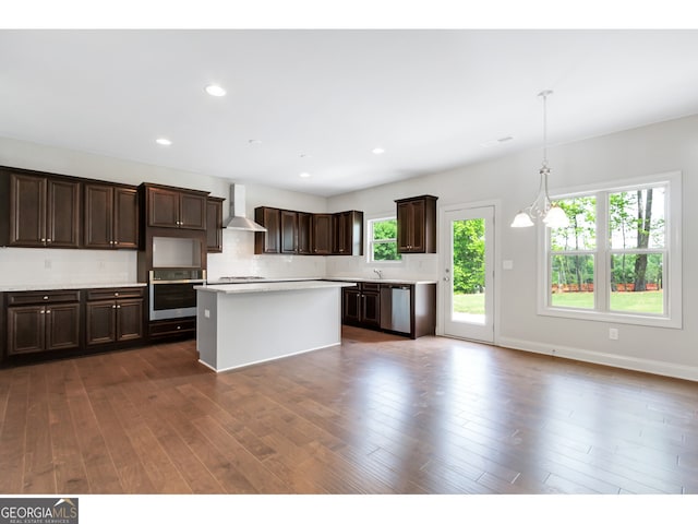 kitchen featuring wall chimney range hood, appliances with stainless steel finishes, decorative light fixtures, a kitchen island, and dark brown cabinets