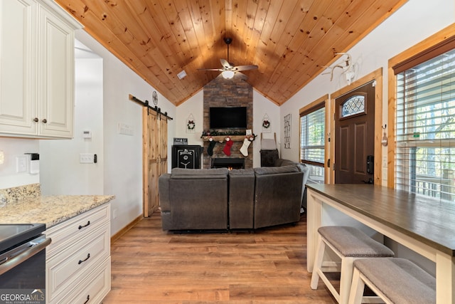 living room featuring vaulted ceiling, ceiling fan, a barn door, light hardwood / wood-style floors, and a stone fireplace