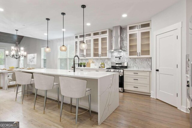 kitchen with light wood-type flooring, wall chimney exhaust hood, a center island with sink, white cabinets, and stainless steel stove