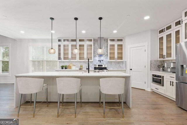 kitchen with a kitchen island with sink, white cabinets, stainless steel appliances, and wall chimney range hood