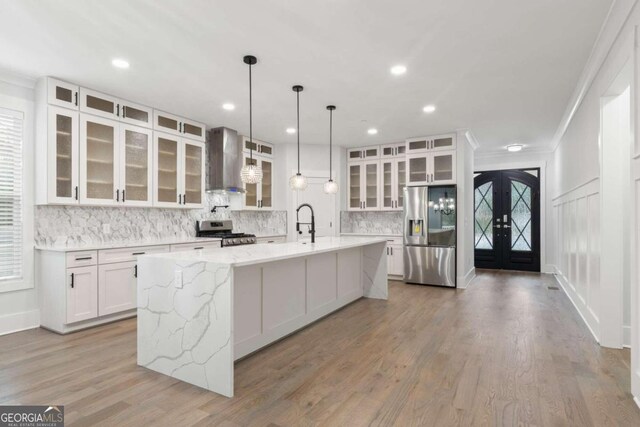 kitchen featuring french doors, wall chimney exhaust hood, stainless steel appliances, a large island with sink, and white cabinets