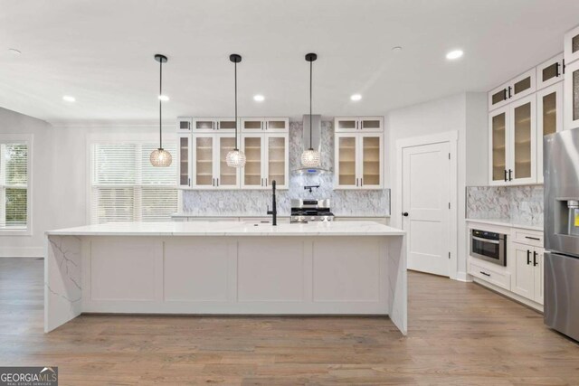 kitchen with appliances with stainless steel finishes, wall chimney exhaust hood, a kitchen island with sink, white cabinets, and light hardwood / wood-style floors