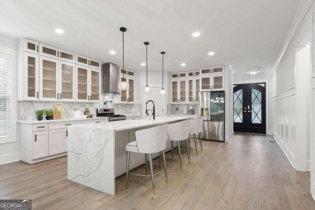 kitchen featuring french doors, stainless steel appliances, wall chimney range hood, a large island with sink, and white cabinets