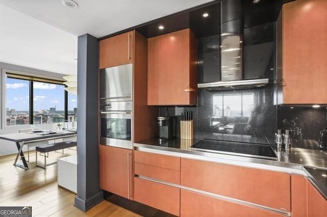 kitchen featuring wall chimney exhaust hood, light wood-type flooring, appliances with stainless steel finishes, and tasteful backsplash