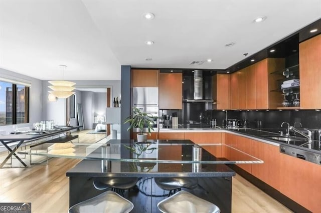 kitchen featuring light wood-type flooring, tasteful backsplash, wall chimney range hood, hanging light fixtures, and a breakfast bar area