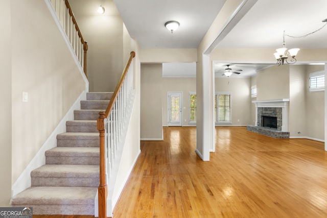 stairs featuring a healthy amount of sunlight, wood-type flooring, ceiling fan with notable chandelier, and a brick fireplace