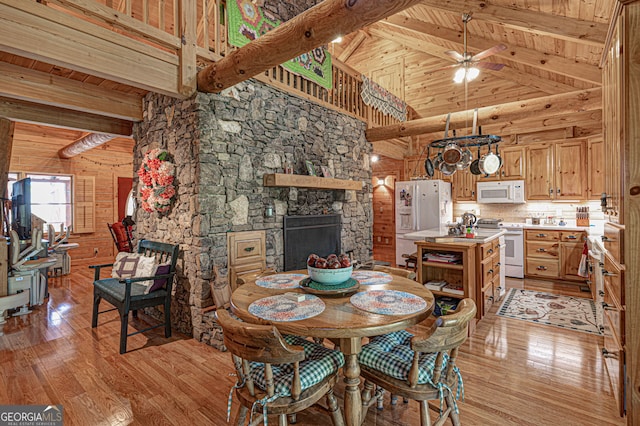 dining space with wooden walls, beam ceiling, high vaulted ceiling, light hardwood / wood-style floors, and a stone fireplace