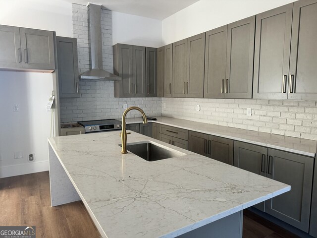 kitchen with a kitchen island with sink, dark wood-type flooring, wall chimney range hood, sink, and light stone countertops