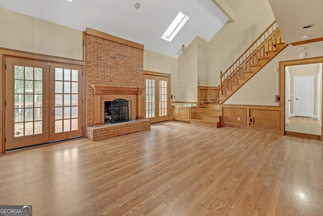 unfurnished living room featuring a skylight, french doors, a brick fireplace, high vaulted ceiling, and light hardwood / wood-style floors