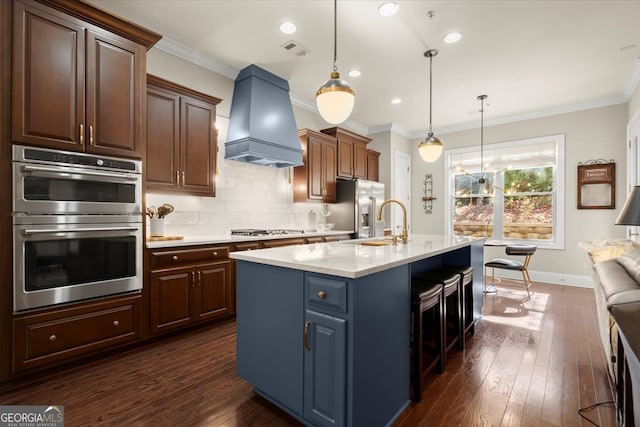 kitchen featuring appliances with stainless steel finishes, custom range hood, dark hardwood / wood-style floors, hanging light fixtures, and an island with sink