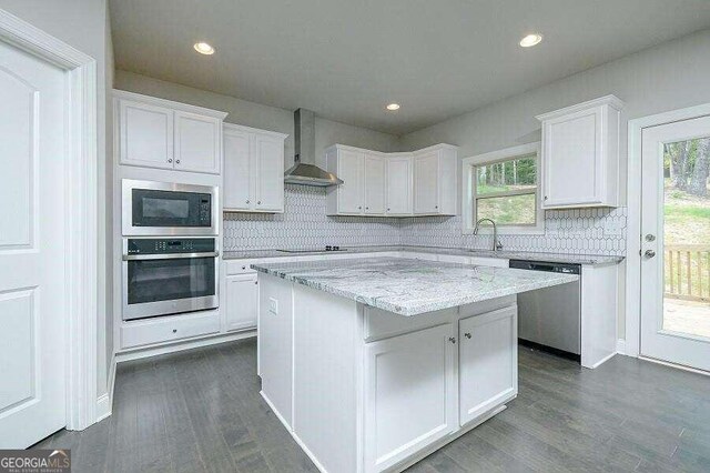 kitchen with wall chimney range hood, dark wood-type flooring, appliances with stainless steel finishes, white cabinetry, and a kitchen island