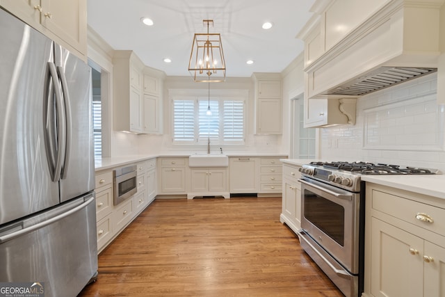 kitchen featuring custom exhaust hood, sink, hanging light fixtures, appliances with stainless steel finishes, and light hardwood / wood-style floors