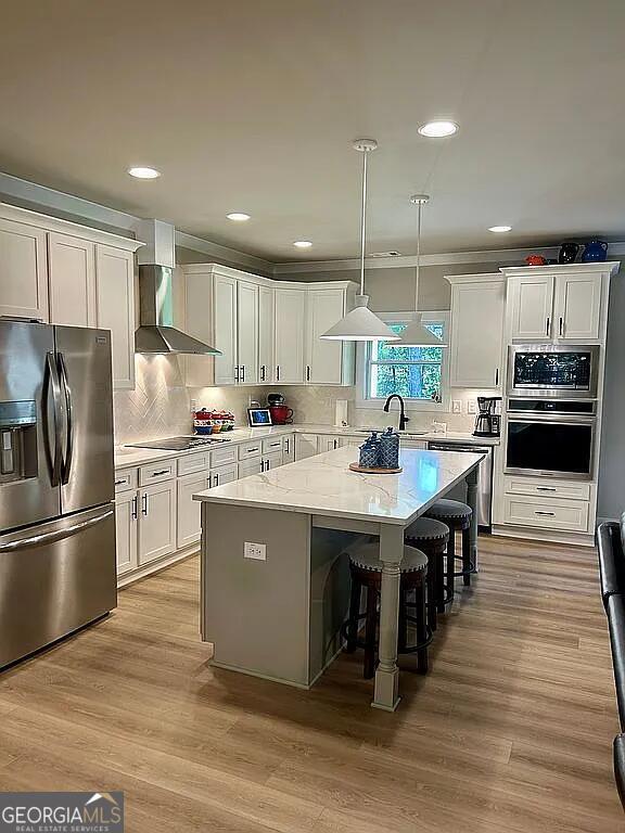 kitchen with white cabinetry, a center island, wall chimney exhaust hood, stainless steel appliances, and decorative light fixtures