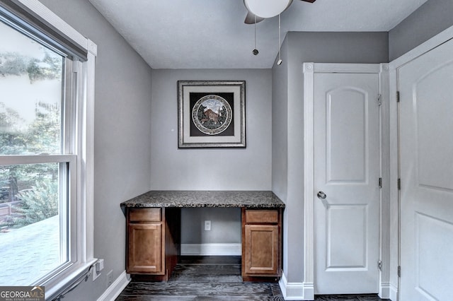 unfurnished office featuring built in desk, plenty of natural light, dark wood-type flooring, and ceiling fan