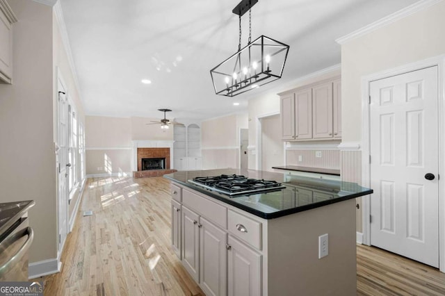 kitchen with light wood-type flooring, a brick fireplace, ceiling fan with notable chandelier, stainless steel gas cooktop, and a kitchen island