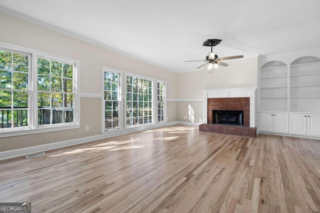 unfurnished living room featuring a brick fireplace, built in shelves, ceiling fan, crown molding, and light hardwood / wood-style flooring