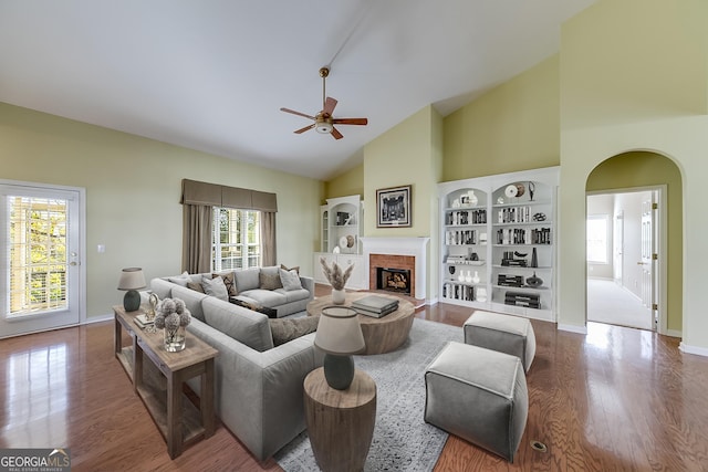 living room featuring built in features, high vaulted ceiling, hardwood / wood-style flooring, ceiling fan, and a brick fireplace