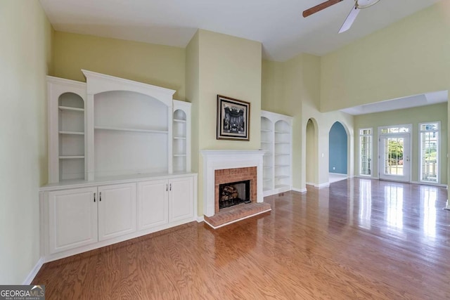 unfurnished living room featuring a brick fireplace, built in shelves, ceiling fan, high vaulted ceiling, and light hardwood / wood-style flooring