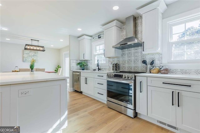 kitchen featuring white cabinets, wall chimney range hood, hanging light fixtures, appliances with stainless steel finishes, and light hardwood / wood-style floors