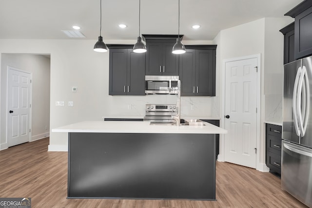 kitchen with a kitchen island with sink, light hardwood / wood-style flooring, decorative light fixtures, and appliances with stainless steel finishes