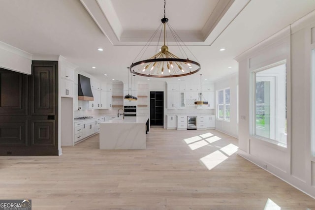 kitchen with white cabinets, decorative light fixtures, an island with sink, a notable chandelier, and custom range hood