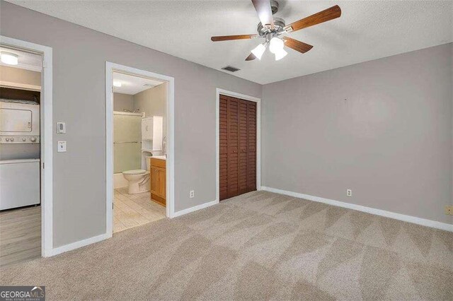 unfurnished bedroom featuring ceiling fan, stacked washing maching and dryer, ensuite bathroom, light carpet, and a closet