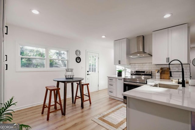 kitchen with stainless steel electric range, sink, light hardwood / wood-style flooring, wall chimney exhaust hood, and white cabinetry