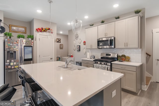 kitchen featuring light wood-type flooring, stainless steel appliances, a kitchen island with sink, sink, and decorative light fixtures