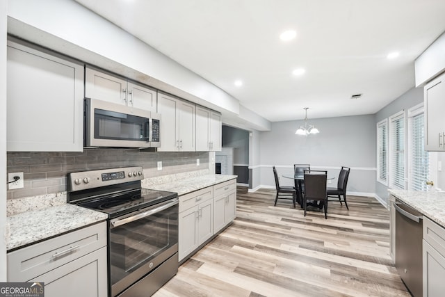 kitchen with light stone countertops, backsplash, stainless steel appliances, an inviting chandelier, and light hardwood / wood-style floors