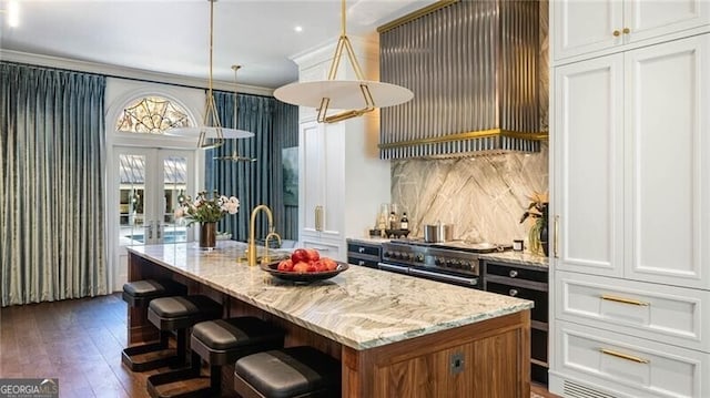 kitchen with an island with sink, decorative light fixtures, white cabinetry, custom exhaust hood, and range with two ovens