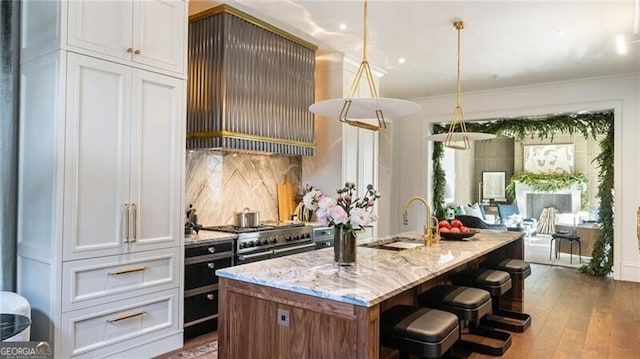 kitchen with an island with sink, white cabinetry, decorative light fixtures, range with two ovens, and wall chimney range hood