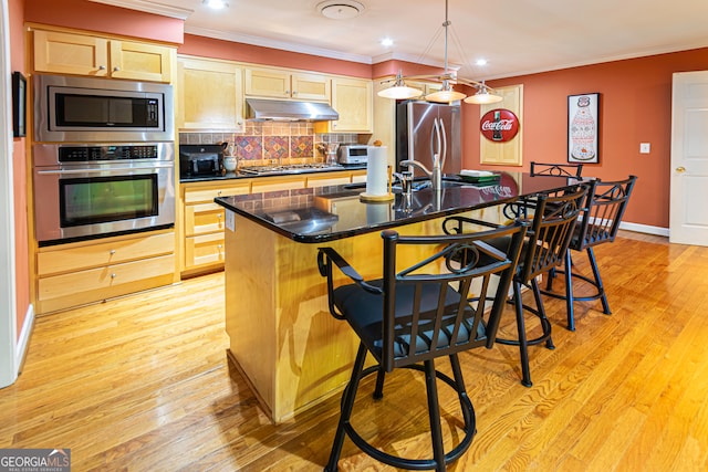 kitchen featuring a center island with sink, light wood-type flooring, light brown cabinetry, appliances with stainless steel finishes, and a breakfast bar area