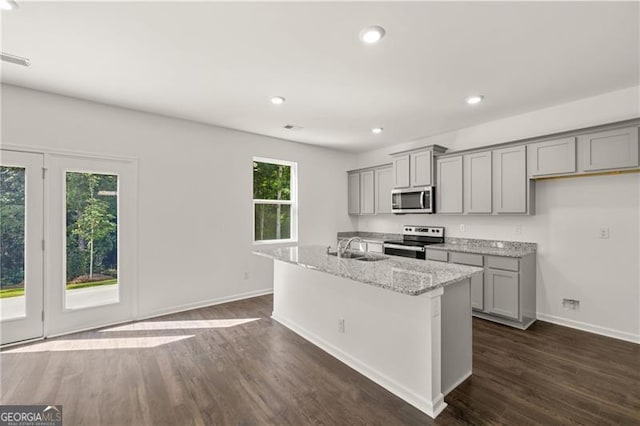 kitchen featuring gray cabinetry, dark wood-type flooring, stainless steel appliances, light stone counters, and a center island with sink