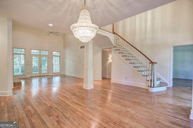 unfurnished living room with french doors, a towering ceiling, decorative columns, ceiling fan with notable chandelier, and wood-type flooring