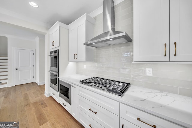 kitchen featuring white cabinets, light wood-style floors, light stone counters, stainless steel appliances, and wall chimney range hood