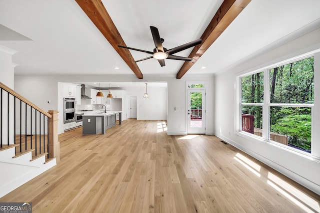 unfurnished living room featuring baseboards, light wood-style flooring, beamed ceiling, stairs, and a sink