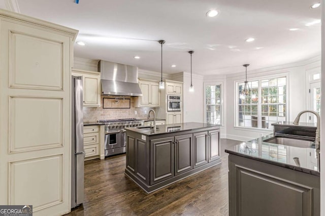 kitchen with sink, hanging light fixtures, wall chimney range hood, cream cabinetry, and appliances with stainless steel finishes