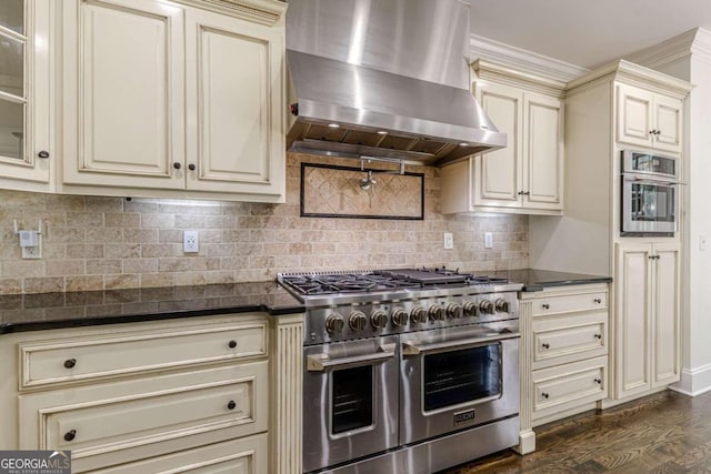 kitchen with wall chimney exhaust hood, dark wood-type flooring, backsplash, cream cabinetry, and appliances with stainless steel finishes