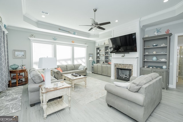 living room featuring ceiling fan, light hardwood / wood-style flooring, crown molding, a tray ceiling, and a fireplace