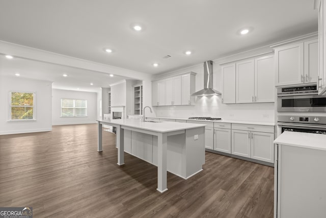 kitchen with appliances with stainless steel finishes, wall chimney exhaust hood, dark wood-type flooring, a center island with sink, and white cabinets